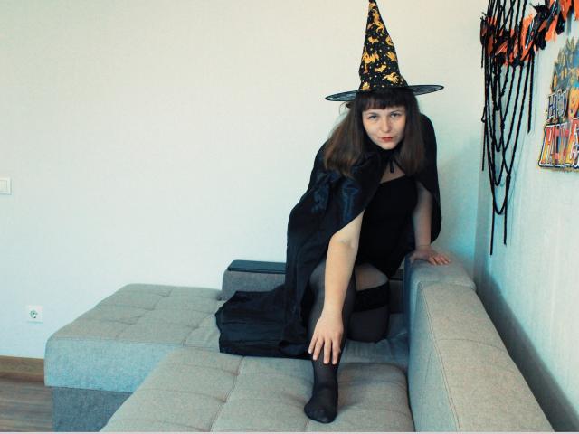 AliceInks with brown hair, wearing a black witch dress, black cape, and yellow-black witch hat, sitting on a grey sectional, one leg up, showing her black pantyhose.