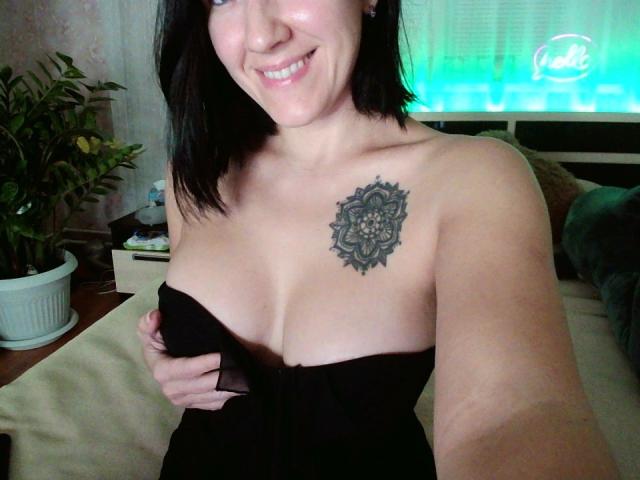 Misstyk with black hair, smiling, showing a large mandala tattoo on her right breast, holding black dress to expose her tits, in a bedroom with green neon light.