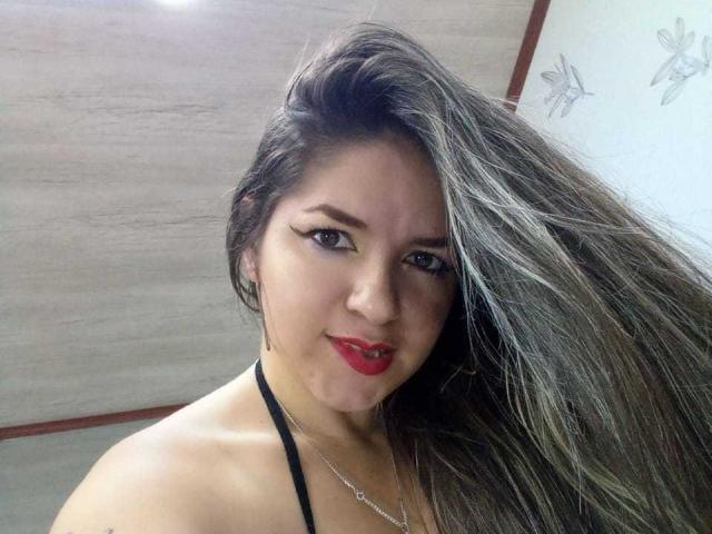 AbbyShiine with long, flowing brown hair, red lipstick, black halter top, and eyeliner, smiling at the camera, showing off her tits. She's wearing a necklace.