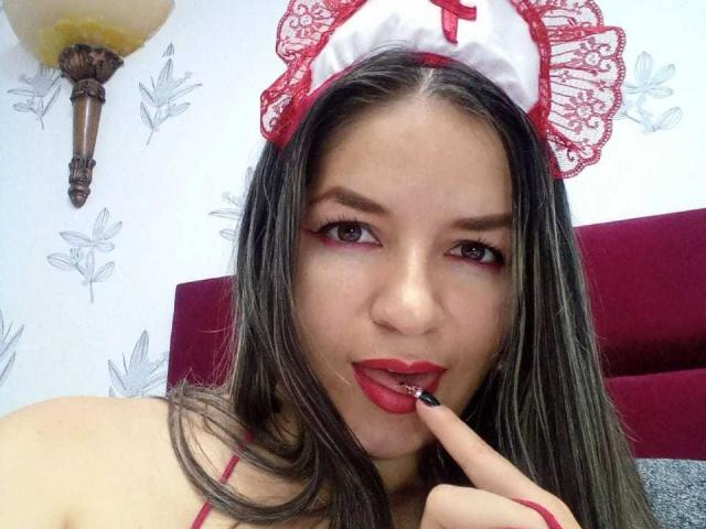 AbbyShiine with long brown hair, red lipstick, and lace nurse cap, biting her red-painted finger, showing her slightly parted red lips and seductive gaze.