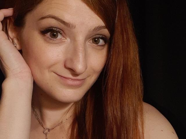 AriaMermaid with red hair, brown eyes, fair skin, and a slight smile, holding her head, wearing a necklace, showing her bare shoulders and a relaxed, cute expression.