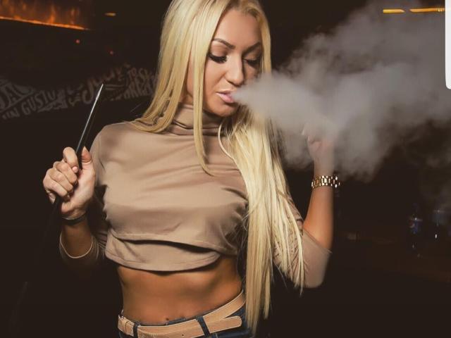 Liliana, con lunghi capelli biondi, tiene in mano un telefono e fuma una sigaretta elettronica, indossa un top corto color pelle e jeans a vita alta, e sta soffiando una densa nuvola di fumo..