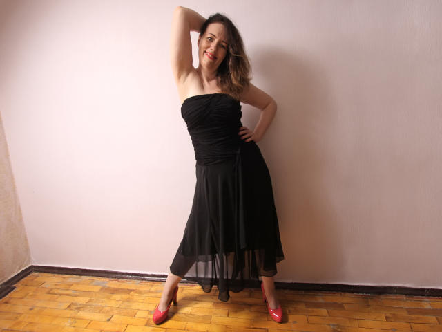 BethShine è in piedi con un braccio dietro la testa, indossa un abito nero senza spalline e scarpe rosa, con i glutei leggermente sollevati, sorridendo e con una mano appoggiata sul fianco..