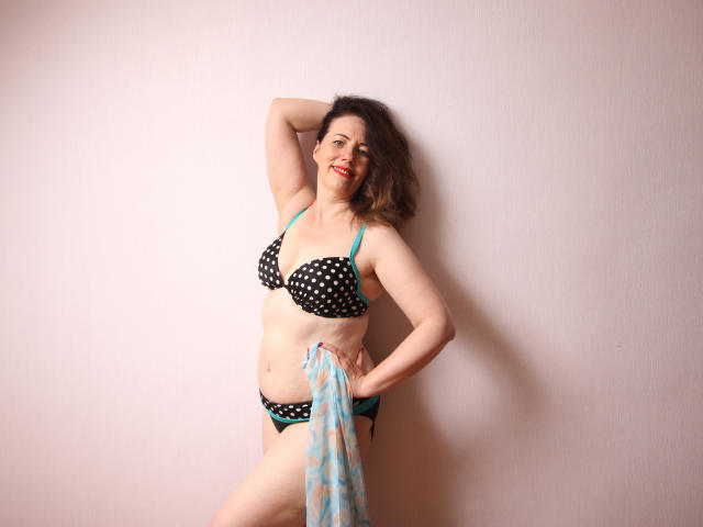 Beth, con un bikini nero e turchese a pois, tiene uno scialle blu trasparente, in piedi di fronte a un muro rosa, sorride con il rossetto rosso, con un braccio alzato, mostrando il seno e i glutei..