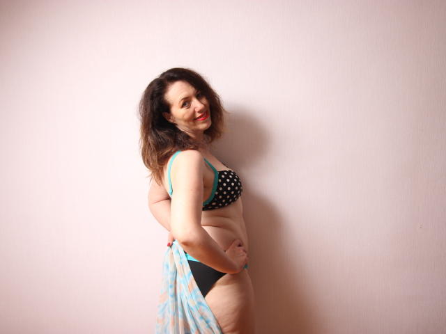 Beth, con capelli marroni medi, indossa un reggiseno nero e azzurro a pois e biancheria intima nera, sorridendo, con le mani sui glutei, uno scialle azzurro a motivi e in piedi contro un muro spoglio..