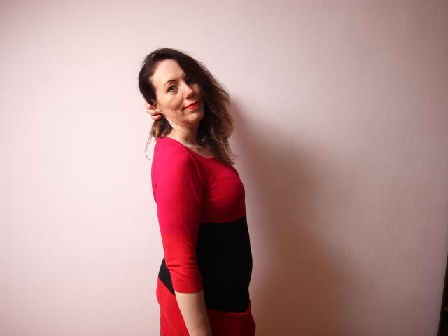 BethShine with wavy brown hair, red and black long-sleeve top, and red pants, standing sideways, smiling at camera, showing her tits and ass subtly.
