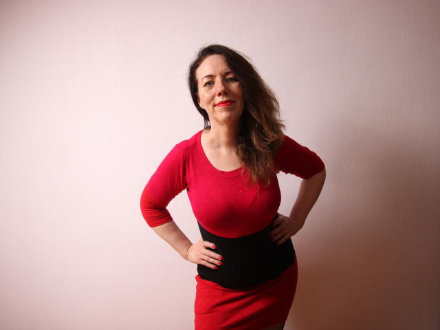 BethShine, com as mãos nos quadris, vestindo um vestido vermelho com faixa preta, cabelos castanhos ondulados, batom vermelho e um sorriso confiante. Seus seios estão levemente visíveis através do vestido..