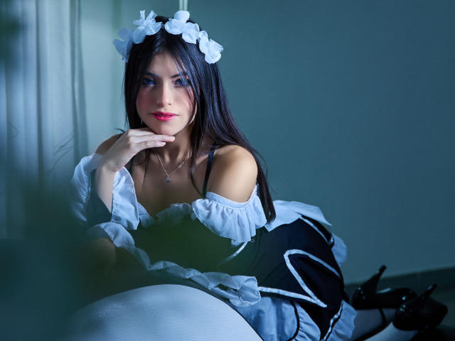 VanityValos mit langen, schwarzen Haaren, die eine weiße Blumenkrone trägt, ein blaues Kleid mit Schulterpartie, das von einer Schulter abrutscht, und schwarze High Heels, sitzt und stützt ihr Kinn auf ihrer Hand, während sie in die Kamera schaut..
