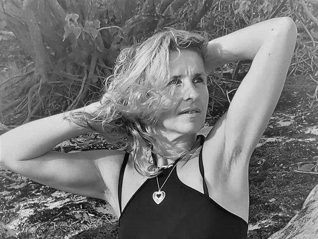 Photo en noir et blanc de TitaLove, avec des cheveux blonds ondulés, les bras levés, montrant les poils des aisselles, portant un collier en forme de cœur, regardant sur le côté, debout à l'extérieur avec un fond de feuilles..