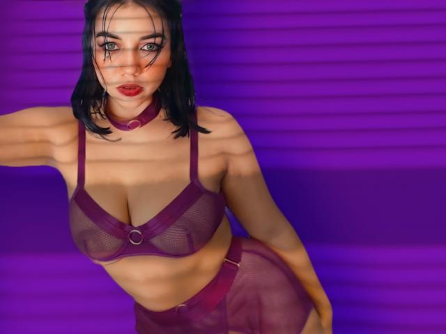 SquirtingCouple with black hair, red lipstick, and purple lingerie, showing her tits in a mesh bra with a ring, and matching panties, against a purple background.
