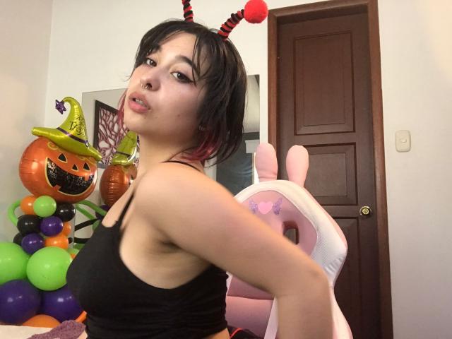 Chloe Lavigne, vestindo uma camiseta preta, uma tiara vermelha e preta com chifres, cabelo curto e escuro, sentada em uma cadeira rosa em formato de coelho, com decoração de Halloween, mostrando seus seios e nádegas. Uma porta marrom ao fundo..