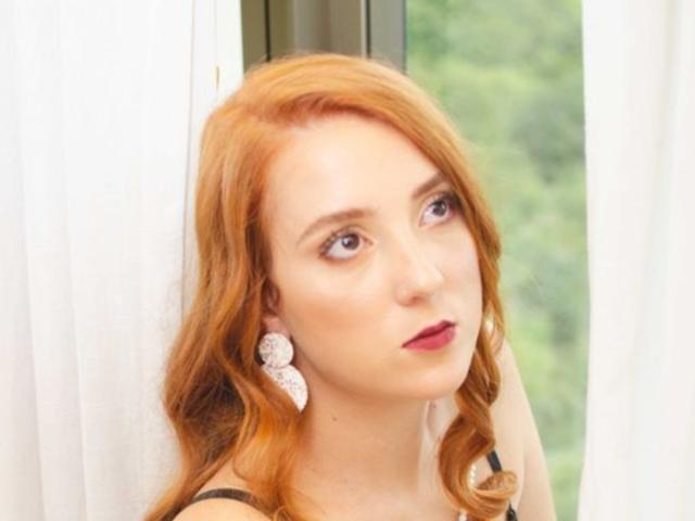 Clarice Mallor, met rood haar, lichte huid en roze lippenstift, draagt een zwarte top en witte oorbellen, en kijkt schuin met een lichte, bedachtzame uitdrukking. De borsten zijn niet zichtbaar..