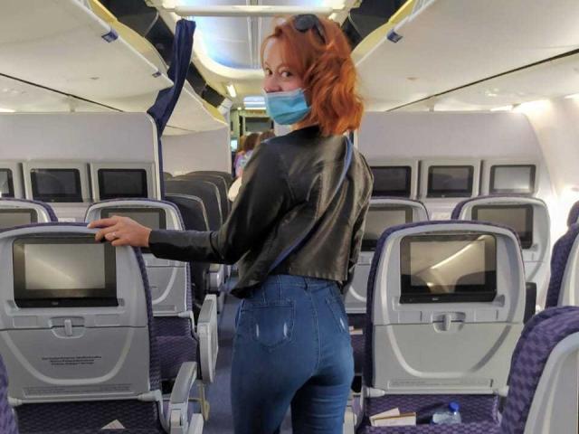 ElizaJones with red hair, wearing a blue mask, black leather jacket, and blue jeans, standing in an airplane aisle, looking back over her shoulder, showing her ass.