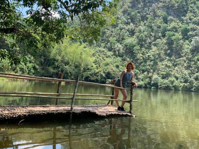 Eliza Jones está de pie en un muelle de madera, vestida con un vestido azul sin mangas, con las nalgas ligeramente levantadas, en un lago verde y tranquilo, rodeado de árboles y ramas..