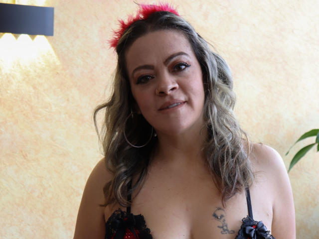 LiliCarina with wavy gray hair, red feather headband, black lace bra showing her tits, small tattoos on right chest, hoop earrings, smiling, indoor light, green plant in background.