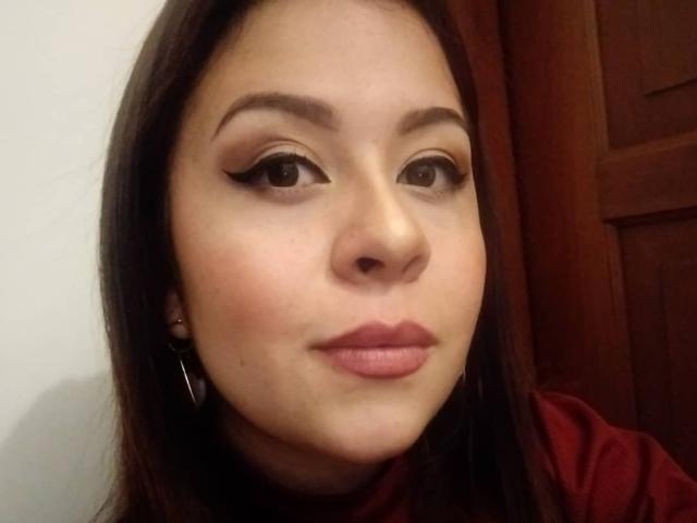 O rosto de Katie Woolf, com olhos castanhos delineados, batom rosa e brincos, vestindo uma camisa vermelha, com cabelo castanho e um leve sorriso..