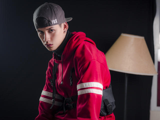JeanKarloLeone draagt een rode hoodie met witte strepen, een zwarte rugzak en een grijze achterstevoren cap. Hij heeft een serieuze uitdrukking, bleke huid en bruine ogen. Er staat een lamp op de achtergrond..