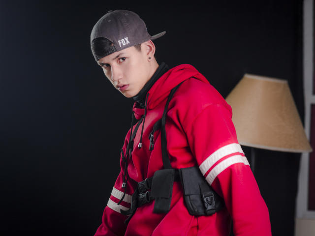 JeanKarloLeone in a red hoodie with white stripes, black harness, and grey backward cap, looking serious, with a nose ring and a slight frown. Lamp in the background.