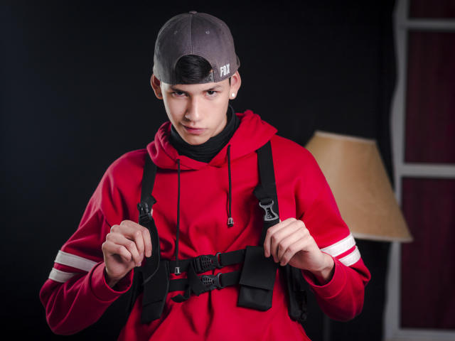 JeanKarloLeone, portant une casquette de baseball grise et un sweat à capuche rouge, serre les sangles noires d'un gilet tactique, avec une expression sérieuse, dans un studio éclairé par une lampe..