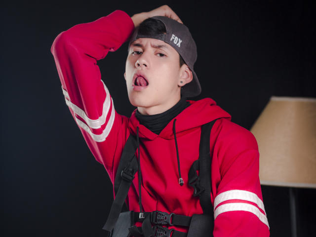 JeanKarloLeone met een zelfverzekerde uitdrukking, tong uit, gekleed in een rode hoodie met witte strepen, zwarte rugzakbanden en een grijze Fox-pet, met zijn rechterarm omhoog..