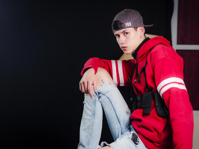 JeanKarloLeone sits with a serious look, wearing a red hoodie, ripped jeans, and a backward grey cap with "FOX" on it, against a dark background.