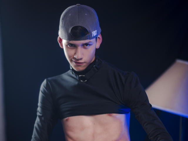 JeanKarloLeone mit flacher Brust, der ein schwarzes Basecap und ein langärmliges Hemd trägt, wobei sein Bauch sichtbar ist. Er hat ein leichtes Lächeln und einen schwarzen Kragen..