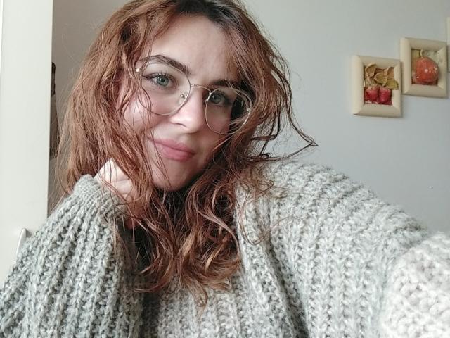 ZoeyFancy mit feuchten, welligen braunen Haaren, die eine Brille trägt und einen dicken, grauen Strickpullover anhat, lächelt leicht und wirkt dabei niedlich und entspannt. Ihre Brüste sind nicht sichtbar..