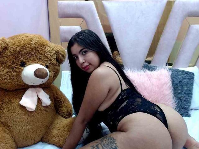 AshleyRoss, met lang zwart haar, draagt een zwarte kanten string, zit op haar knieën, naast een bruin knuffelbeer, en toont haar achterwerk en een tatoeage op haar rechter dij..