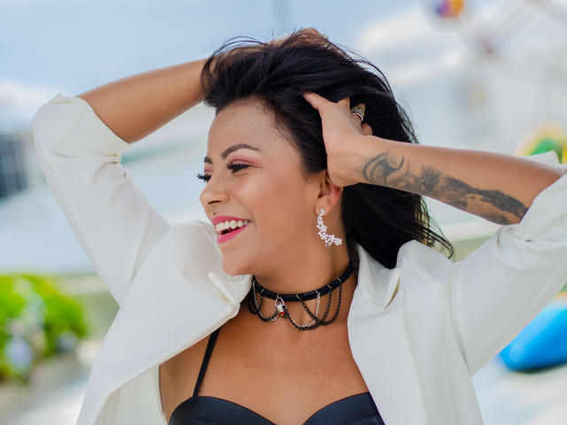 Amberv with dark hair, wearing a white shirt, black strap top, and choker, smiling, hands in hair, tattoo on arm, earrings, pink lipstick, sunny background, looking to the side.