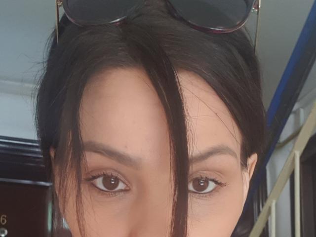 JoliDanielle's face with dark brown eyes, subtle eyeliner, and straight black hair, partially covering her forehead, wearing sunglasses on top. Her skin is light brown and smooth.