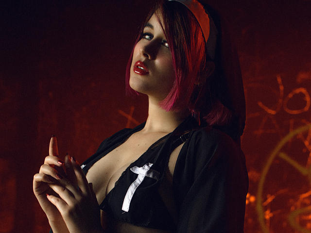 MaraMaison avec du rouge à lèvres, des cheveux roses et bruns, un haut noir transparent, une croix blanche, montrant les seins, les mains jointes, regardant vers le haut, fond rouge foncé, ombragé, sexy, confiante..