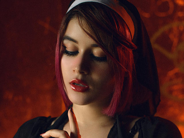 MaraMaison con rossetto rosso, occhi chiusi e caschetto rosa, che indossa un abito da suora, toccandosi il mento con un dito, con un trucco scuro e un'espressione sexy e sensuale..