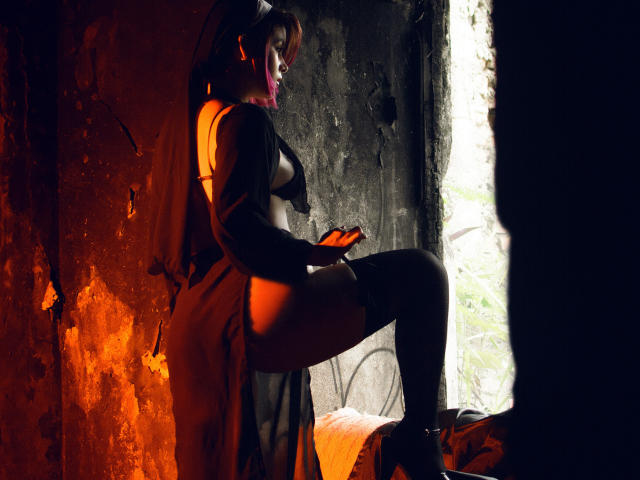 MaraMaison mit freiliegenden Brüsten und Gesäß, mit lila Lippenstift, trägt einen durchsichtigen schwarzen Bademantel und hohe Socken, steht in einem dunklen, orange beleuchteten Raum und hält ein orangefarbenes Telefon..