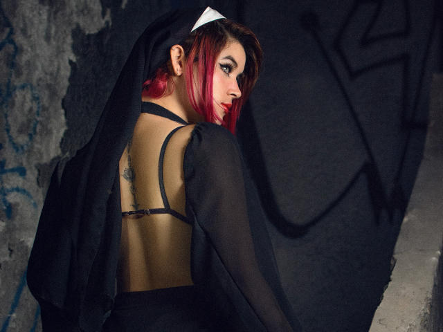 MaraMaison, con el cabello teñido de rojo, un vestido negro transparente y un tocado blanco, mostrando su tatuaje en la espalda y las tiras de su sujetador negro, mirando por encima del hombro con ojos seductores..