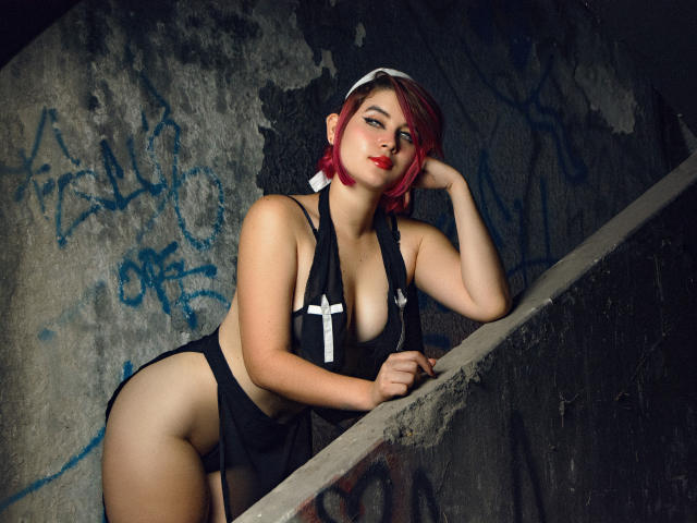MaraMaison, con cabello rojo, lencería negra y un crucifijo, apoyada en una pared, mostrando sus pechos y glúteos, luciendo sexy, en un entorno urbano y descuidado, cubierto de grafitis..