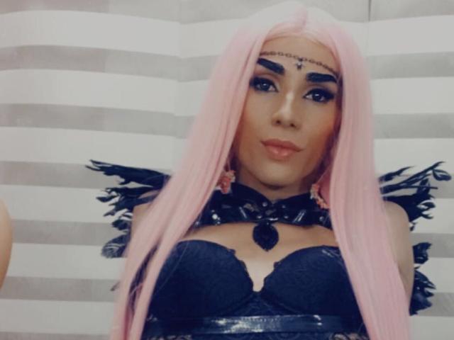CrazyManHot with pink hair, dark makeup, and a black lace bustier, showing off her tits and black winged shoulders, looking hot and sexy.