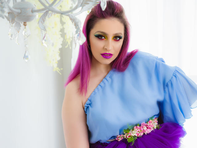 Agaa, met roze haar, een blauwe jurk, een paarse rok en roze bloemen, heeft gele en roze oogmake-up en roze lippenstift, en leunt tegen een witte muur met een kroonluchter..