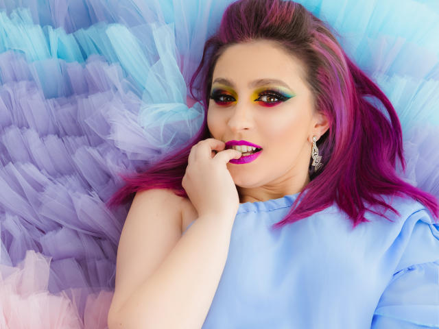 Agaa a les cheveux rose et violet, un maquillage des yeux jaune-vert et noir, du rouge à lèvres violet, et porte un haut bleu. Son doigt est près de sa bouche ouverte, et elle a l'air séduisante..