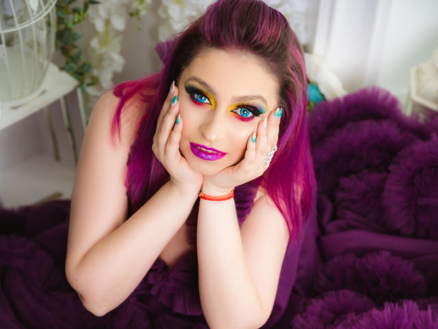 Agaa with bright purple hair, blue and yellow eye makeup, and purple lipstick, hands on her face, blue nails, orange bracelet, and ring, sitting on a purple fluffy blanket.