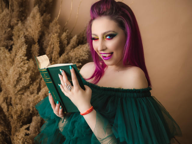 Agaa, con i capelli viola brillante, un abito verde a spalle scoperte e un trucco colorato, tiene in mano un libro verde, sorride, mostrando i denti bianchi, con le unghie color acquamarina e un braccialetto rosso..