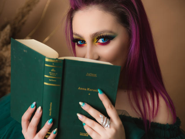 Agaa mit lila Haaren, blauem und gelbem Augen-Make-up, liest ein grünes Buch, hat türkis gefärbte Nägel, trägt ein Oberteil mit einem Schulterauschnitt und zeigt einen Ring an ihrer rechten Hand..