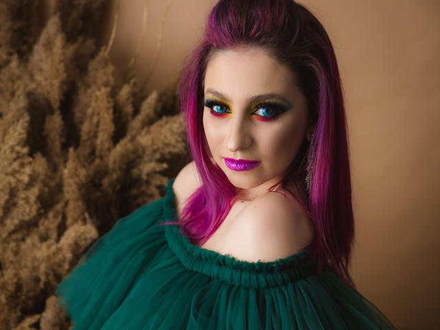 Agaa, com cabelo roxo, vestido verde com um ombro à mostra e batom roxo, tem sombra colorida e um sorriso adorável. Os seios estão parcialmente visíveis. Ela está olhando para cima com uma expressão fofa..