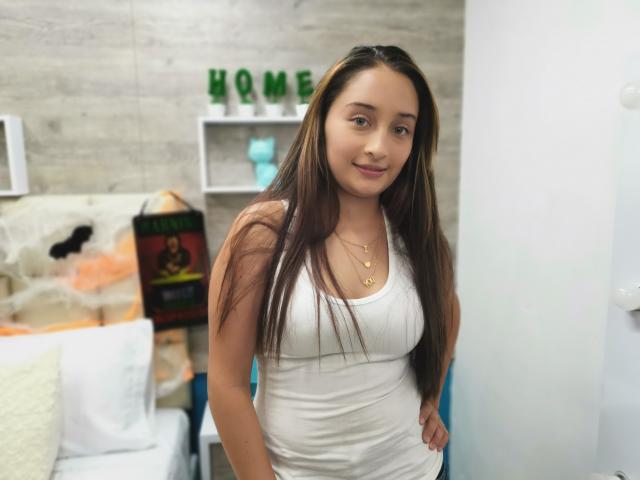 BeckyMils with long brown hair, wearing a white tank top, standing, hands on hips, smiling, small gold necklace, cute, slender, small tits, soft light, casual room.