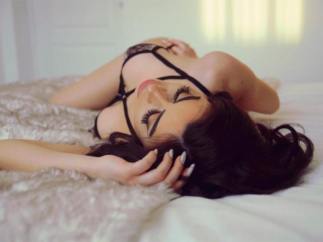 LauraineX with dark hair, lying on a fluffy bed, wearing black lingerie, eyes closed, hand in hair, long eyelashes, pink lips, white nails, soft light, sensual pose.
