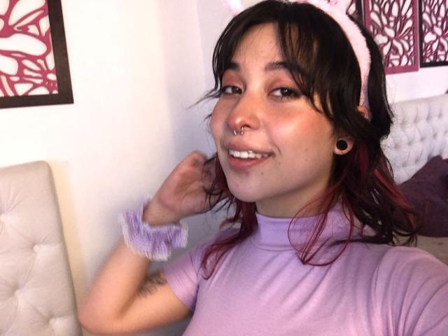 ChloeLavigne with dark brown hair, pink highlights, white headband, purple shirt, nose ring, ear gauges, smiling, wrist scrunchie, holding hair, white tufted chair, wall art in background.