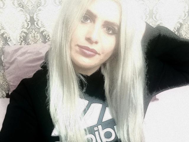 EveEveline with long white hair, wearing a black Adidas shirt, has pink lipstick, light skin, and a slight smile, sitting with one hand behind her head.