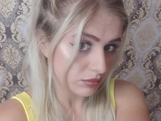 Eveline, con cabello rubio, ojos verdes y un pequeño piercing en la nariz, viste una blusa amarilla, tiene una ligera expresión de ceño fruncido y un maquillaje sutil con delineador de ojos..