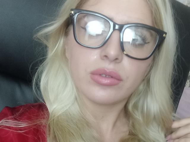 EveEveline a les cheveux blonds, des lunettes à monture noire épaisse, des lèvres roses brillantes et une chemise rouge. Son visage est légèrement brillant, reflétant une source de lumière dans ses lunettes..