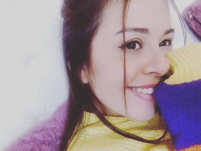 Katie Woolf, met een schattende glimlach, bruine ogen en een gele top, houdt een pluizig speelgoed in paars en geel vlakbij haar neus. Haar bruine haar is iets rommelig..