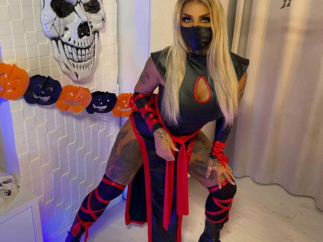 BarbaraBae con lunghi capelli biondi, maschera nera, abito da ninja grigio, fiocco rosso, bracciali neri, gambe tatuate, in posizione accovacciata, teschio di Halloween, zucche intagliate arancioni e nere, pavimento bianco..